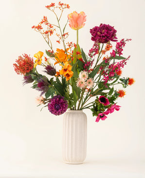 A white, ribbed ceramic vase displays the Magnus di Lambo silk artificial flower bouquet - an arrangement of vibrant orange, pink, purple and yellow artificial flowers with green leaves against a light background.