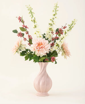 A pink ceramic vase contains the Morning Whisper silk artificial flower bouquet, with pale pink dahlias, white snapdragons, green leaves and bunches of small red berries, against a plain off-white background.