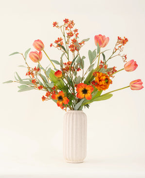 The Orange Glory silk artificial flower bouquet, with orange tulips, poppies and green leaves, is beautifully arranged in a white textured vase against a plain light background.