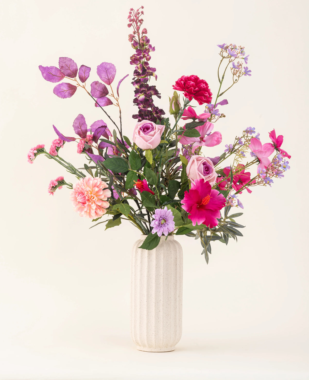 A white textured vase contains the Purple Dawn silk artificial flower bouquet in pink, purple and lavender shades with green foliage, against a light, neutral background.