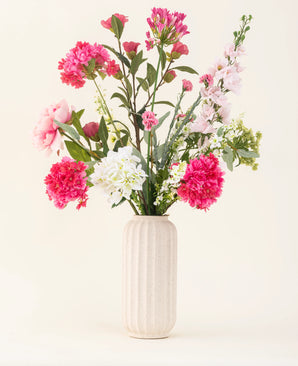 A white textured vase shows the Blush Breeze silk artificial flower bouquet - pink, white and light purple silk flowers with green leaves - against a plain light background.