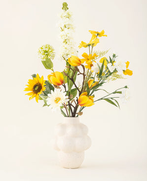 The Sun Glow silk artificial flower bouquet with yellow tulips, sunflowers, daisies and green leaves is placed in a white structured vase against a plain off-white background.
