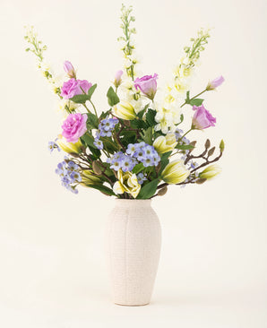 In a cream-colored vase is a silk artificial flower bouquet Magnolia Glow with purple, white and yellow flowers - delphiniums, lisianthus, lilies and forget-me-nots against a plain light background.