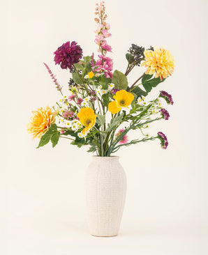 The Daisy Daydream silk artificial flower bouquet features a white textured vase and a vibrant mix of yellow, pink, purple and white flowers with green leaves, adding lasting beauty to any plain light background.