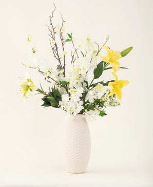 A vase with a white structure shows the elegance of the Sunkissed Ivory silk artificial flower bouquet, with yellow lilies, white flowers and green leaves against a plain light background.