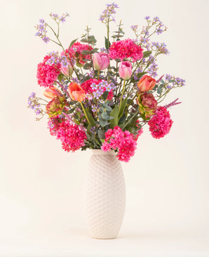 A vase with a white structure and the Soft Spring silk artificial flower bouquet with bright pink flowers, orange tulips and purple-blue blossoms brings spring into your home against a plain light background.