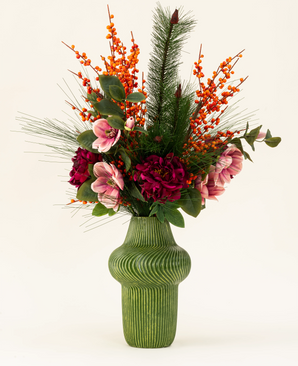 The Silk artificial flower bouquet Cozy Winter presents a green ribbed vase with dark red and pink flowers, orange berries, pine branches and green leaves - perfect as an autumn-winter decoration against a light background.