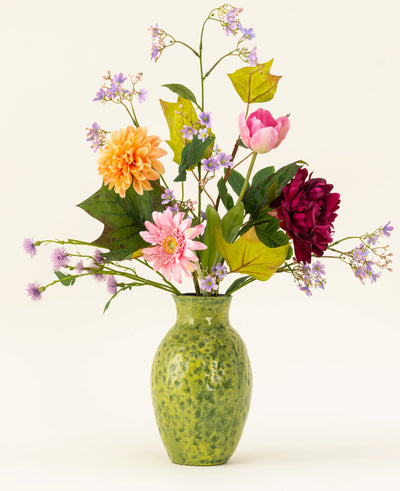 A green speckled vase shows the Silk artificial flower bouquet Love in Bloom, with pink, orange and dark red flowers with purple and green foliage against a light background.