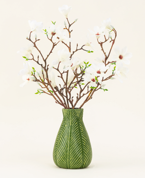 A green vase shows the Silk artificial flower bouquet Magnolia white, with several white magnolia branches with small green leaves, against a plain light background.