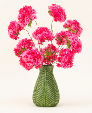 A green vase with leaf motif displays the Artificial Silk Flower Bouquet Cherry Blossom Pink - bright pink, fluffy silk cherry blossoms with green leaves - against a plain light background.