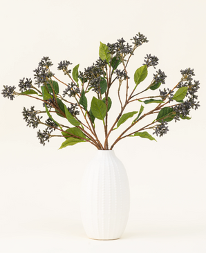 A Silk artificial flower bouquet Japanese Berry Steel Blue with long stems, dark purple berries and steel leaves stands in a vase with white texture against a plain light background.