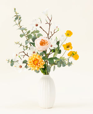The Fields of Joy silk artificial flower bouquet features yellow, white and peach flowers with green eucalyptus in a white textured vase, beautifully arranged against a light background.