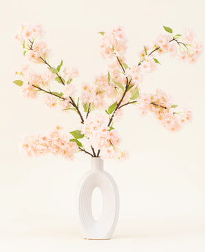 A white ceramic vase with a wide oval opening shows several green-leaved Silk artificial flower bouquet Cherry blossom light pink branches against a simple light background.