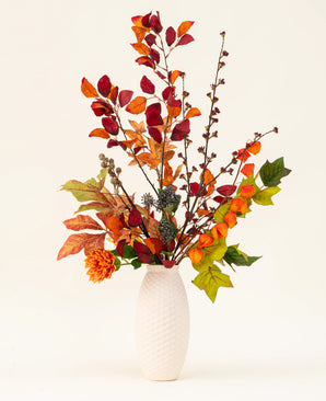 A white textured vase showcases the Chestnut Charm Silk Artificial Flower Bouquet, featuring autumn leaves, orange flowers and green foliage - an elegant artificial arrangement against a plain light background.