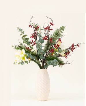 The Silk artificial flower bouquet Midwinter Dream has green leaves, pine branches, red berries and white flowers in a vase with a white structure - perfect for a serene winter bouquet against a plain light background.