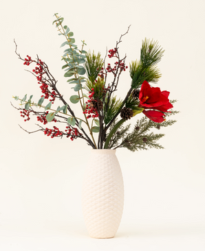 The Winterberry Bliss silk artificial flower bouquet features red berries, pine branches, eucalyptus, red amaryllis and pine cones in a white textured vase - ideal for Christmas decoration or to display against a light background.