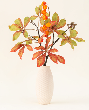 The Silk artificial flower bouquet Autumn Flair has a vase with white structure and lantern branch, orange lantern-shaped flowers and green-red leaves, which form a stylish autumn display against a plain light background.