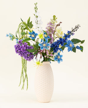 A white structured vase contains the Midnight Valley silk artificial flower bouquet, featuring vibrant blue, purple and yellow Real Touch flowers with several green leaves against a light background.