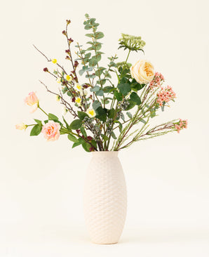 A white textured vase showcases the Blush Blossom silk artificial flower bouquet, featuring real flowers - pale pink roses, yellow flowers and green foliage - against a plain light background.