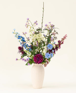A white vase with the Purple Rain silk artificial flower bouquet shows vibrant purple and white artificial flowers and green leaves, an example of Dutch craftsmanship against a light background.