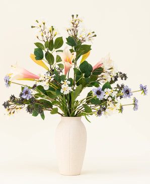 The Blush & Bloom silk artificial flower bouquet - with pink lilies, white and purple flowers and lush green leaves - stands in a white vase against a light background.