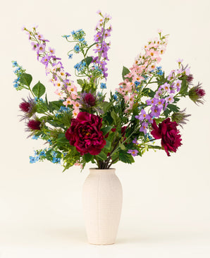 The Moonlit Meadow silk artificial flower bouquet - red peonies, pink and purple delphiniums, blue flowers, green leaves and thistle-like flowers - stands in a vase with a white structure against a plain, light background.