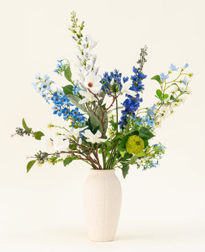 The Cool Breeze silk artificial flower bouquet, with white lilies, blue larkspur and green foliage in a cream-colored vase, stands out against a plain background for a refreshing touch.