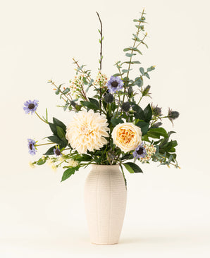 A white ceramic vase contains the Petal Poetry silk artificial flower bouquet, with cream-colored dahlias and roses, light purple flowers, green thistles and all kinds of greenery against a plain light background.