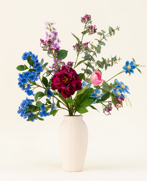 A white textured vase showcases the Painted Petals silk artificial flower bouquet, featuring vibrant blue, purple, pink and red silk flowers with green leaves and eucalyptus branches against a plain light background.