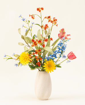 The Lemonade Garden silk artificial flower bouquet features real-tinted sunflowers, pink tulips, blue delphiniums, orange blossoms and green foliage in a beige vase against a plain light background.