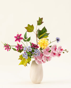 The Aurora Garden silk artificial flower bouquet features real touch flowers in pink, yellow, blue and purple with green leaves, arranged in a white ceramic vase against a plain light background.