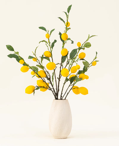 A white textured vase shows the Lemons silk artificial flower bouquet, with green leaves and vibrant yellow lemons against a light background.