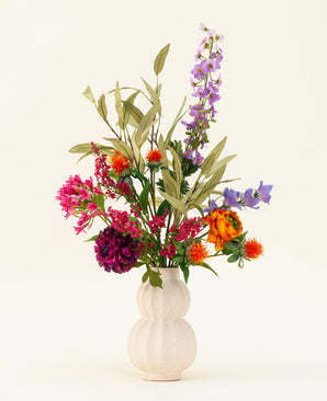 A white, textured vase showcases the Colorful Svenski silk artificial flower bouquet, featuring vibrant purple, orange, pink and blue artificial flowers with green leaves that brighten up any space against a simple light background.