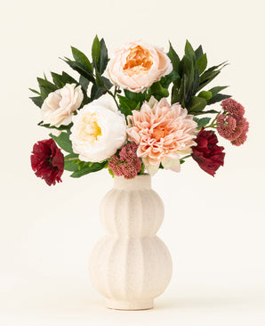 A cream-colored ceramic vase with structure shows the Jewel Bloom silk artificial flower bouquet with pink, white and dark red Real Touch flowers and green leaves against a plain light background.