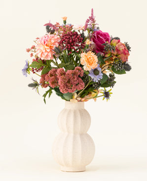 In a white, curved ceramic vase is the Pretty Pastel silk artificial flower bouquet - a colorful mix of sustainable flowers such as pink roses, dahlias, purple and orange flowers and greenery - beautifully arranged against a light background.