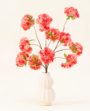 The Silk Artificial Flowers Bouquet Cherry Blossom Coral features vibrant pink and coral artificial cherry blossoms with green leaves, elegantly arranged in a white sculpted vase against a light background.