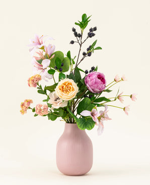 The Touch of Cloudlight silk artificial flower bouquet, presented in a pink ceramic vase, contains pink peonies, yellow roses, small blossoms, greenery and branches with dark berries against a light background.