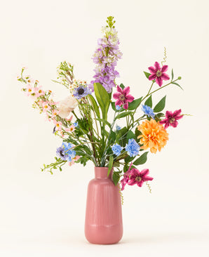 The Silk artificial flower bouquet Bloom & Glow has pink, purple, blue, orange, white and yellow flowers with green leaves in a pink ceramic vase against a light background.