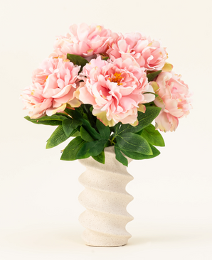 The Silk artificial flower bouquet Peony Soft Pink features large soft pink peonies and green leaves in a wavy, cream-colored ceramic vase, beautifully displayed against a plain light background.