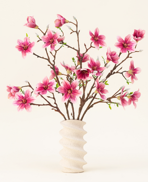 The Silk artificial flower bouquet Magnolia pink is arranged in a wavy white ceramic vase with green buds, creating an elegant display against a light background.