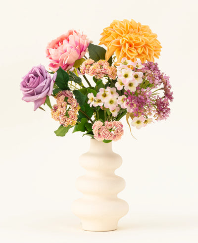 A white corrugated ceramic vase displays the Peach Delight silk artificial flower bouquet, featuring a yellow dahlia, pink peony, purple rose and several small white and pink flowers against a simple light background.