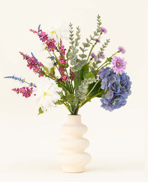 A cream-colored wavy vase contains the Lavender Kiss silk artificial flower bouquet, featuring white dahlias, lilac artificial flowers, purple hydrangeas, pink and purple flowers and green foliage against a plain light background.