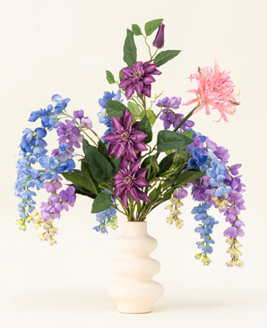 A white wavy vase displays the Indigo Dreams silk artificial flower bouquet - an arrangement of purple, blue and pink artificial flowers with green leaves - against a plain light background.
