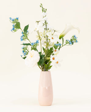 The light pink vase contains the Whispers of White silk artificial flower bouquet, with white flowers, blue blossoms and green foliage against a plain, light background.