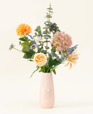 The Earth & Petal silk artificial flower bouquet is a pastel pink vase with lifelike silk flowers in orange, pink and yellow, plus green and purple accents, depicted against a light background.