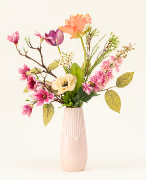 The Spring Awakening silk artificial flower bouquet, with pink magnolias, light yellow poppies and orange and purple flowers in a tall light pink ceramic vase, emphasizes Dutch craftsmanship against a plain white background.