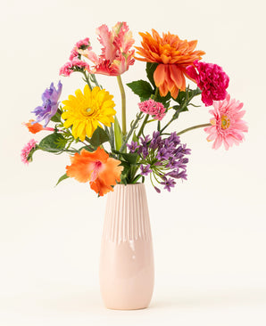 A pale pink ceramic vase contains the Candy Petals silk artificial flower bouquet with vibrant orange, yellow, pink and purple silk flowers against a plain off-white background.