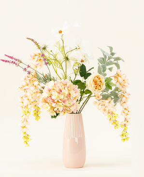 A pastel pink ceramic vase showcases the Blushing Beauty silk artificial flower bouquet, featuring softly colored hydrangeas, roses and assorted greenery, beautifully arranged against a pale neutral background.