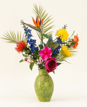 A green, speckled vase shows the Silk artificial flower bouquet Hibiscus Fiesta - with pink, red and yellow flowers, blue delphiniums, orange birds of paradise and green palm leaves - against a plain light background.