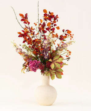 The Golden Autumn silk artificial flower bouquet has an artistic mix of autumn leaves and red, orange and purple flowers in a white ceramic vase, against a light background.